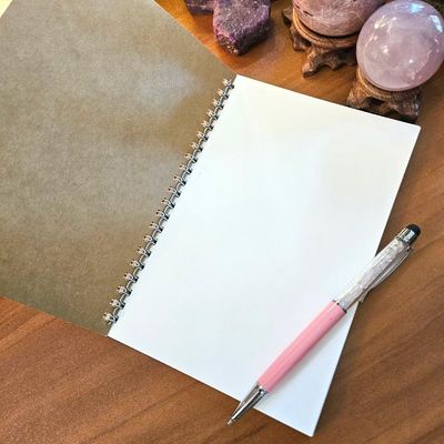 Open spiral‑bound journal with blank white pages on a wooden surface, with a pen and crystals arranged nearby