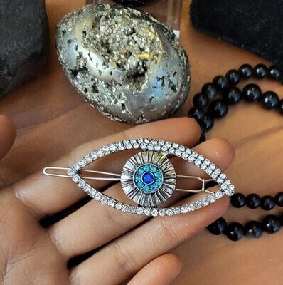 Silver eye‑shaped hair clip with rhinestones and blue gemstones, held near crystals