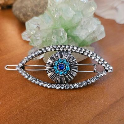 Silver eye‑shaped hair clip with rhinestones and blue gemstones, placed on a wooden surface near a green crystal cluster.
