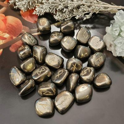 Polished Pyrite rune stones on a dark surface, surrounded by dried herbs and light green crystals