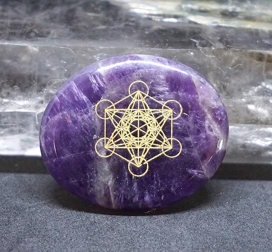 Amethyst palm stone with a gold Metatron engraving placed on a black surface with a crystal in the background.