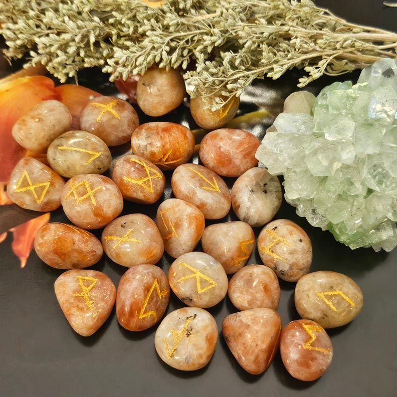 Polished Sunstone rune stones on a dark surface, surrounded by dried herbs and light green crystals
