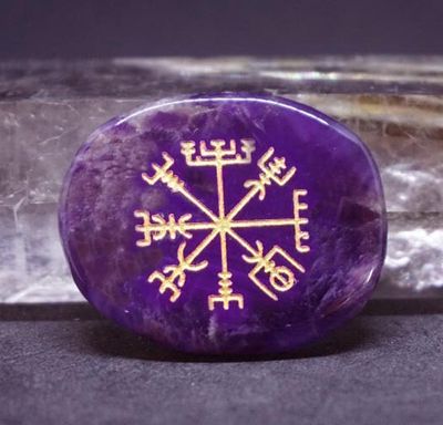 Amethyst palm stone with a gold Vegvisir engraving placed on a black surface with a crystal in the background.