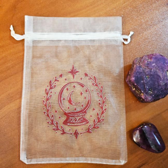 White organza bag with red crystal ball surrounded by moons, stars, and leafy branches.