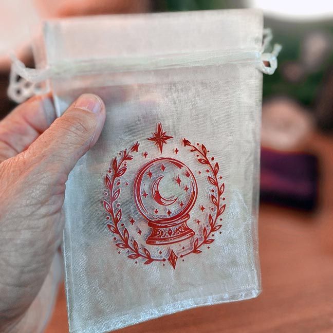 White organza bag with red crystal ball surrounded by moons, stars, and leafy branches.