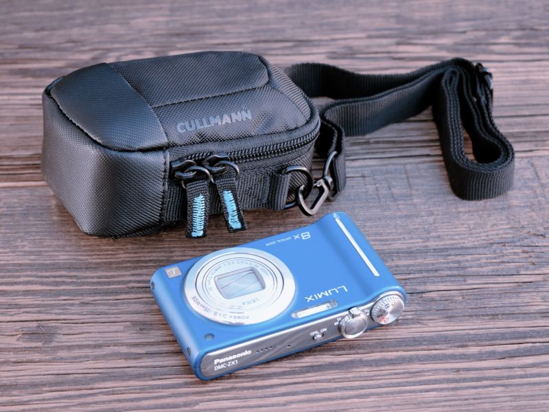 Kompakte schwarze Cullmann Kameratasche mit Schultergurt auf einer Holzoberfläche neben einer blauen Panasonic Lumix DMC-ZX1 Kamera.