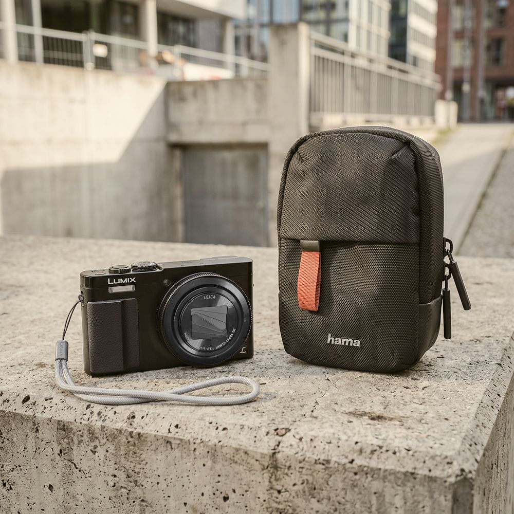 Schwarze kompakte Hama Fototasche mit Gürtelschlaufe auf einem Betonsockel neben einer Panasonic Lumix DMC-TZ71 Kamera, ideal auch für Panasonic Lumix DC-TZ202.