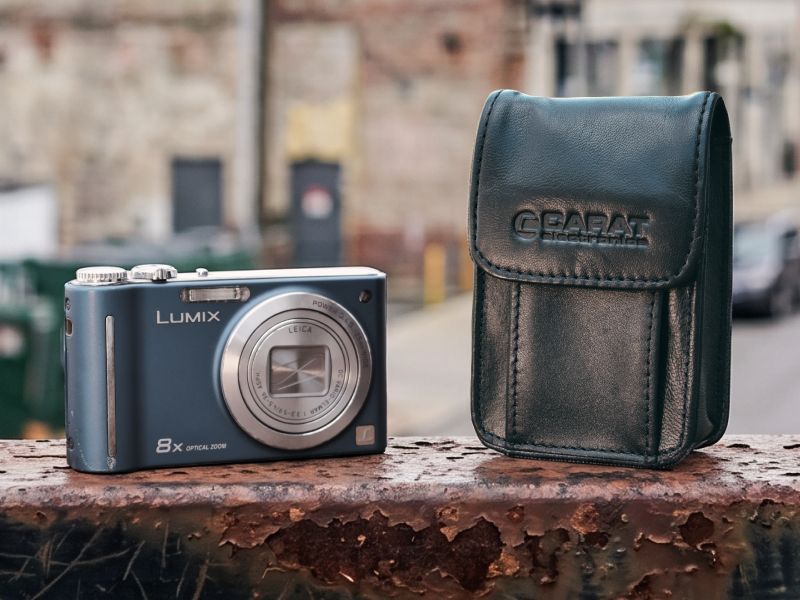 Schwarze Carat Leder-Kameratasche mit Gürtelschlaufe neben einer blauen Panasonic Lumix DMC-ZX1 Kompaktkamera auf einem verrosteten Geländer.