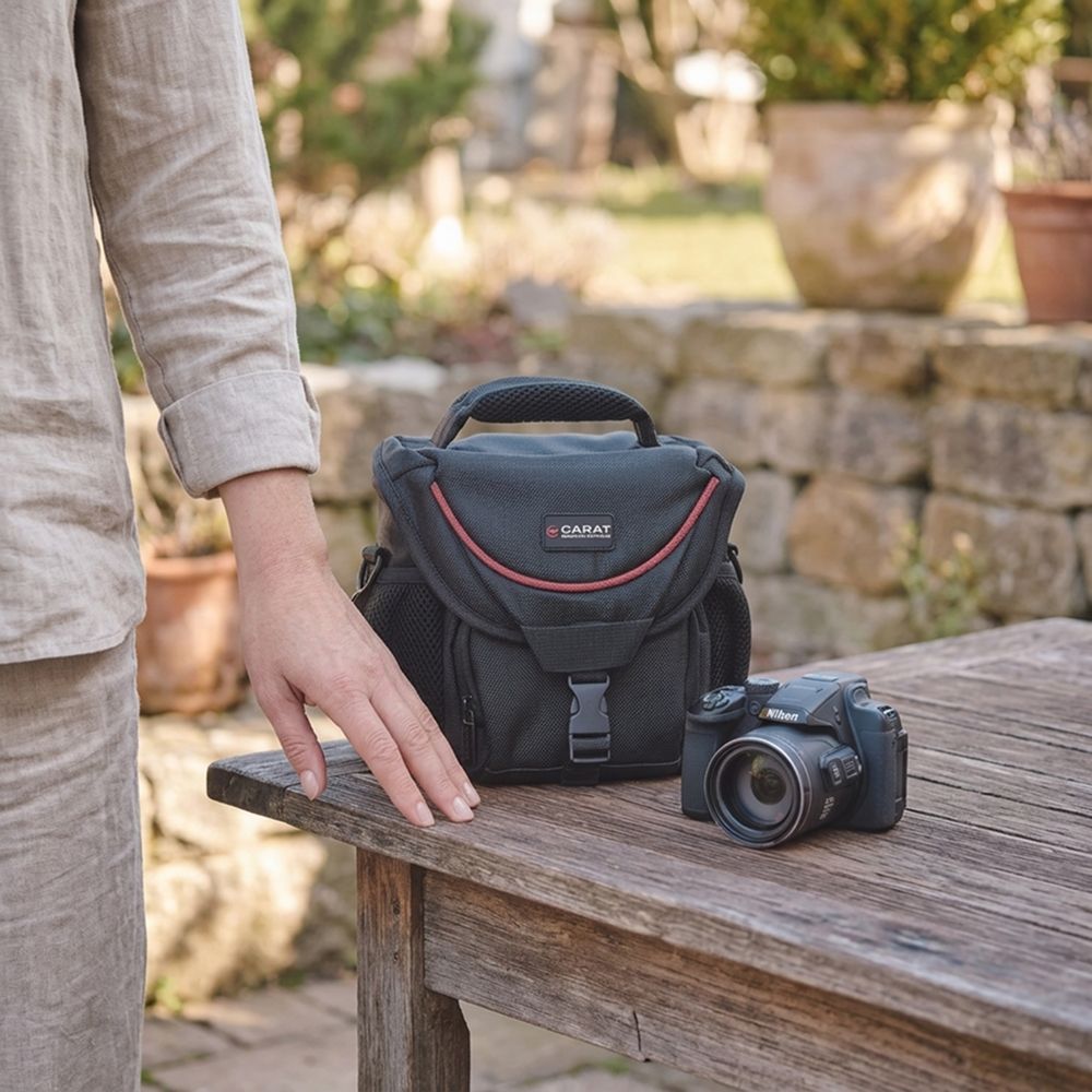 Schwarze Carat Foto-Schultertasche mit Tragegriff auf einem Holztisch neben einer Nikon B700 Bridgekamera, ideal auch für Canon EOS R50.