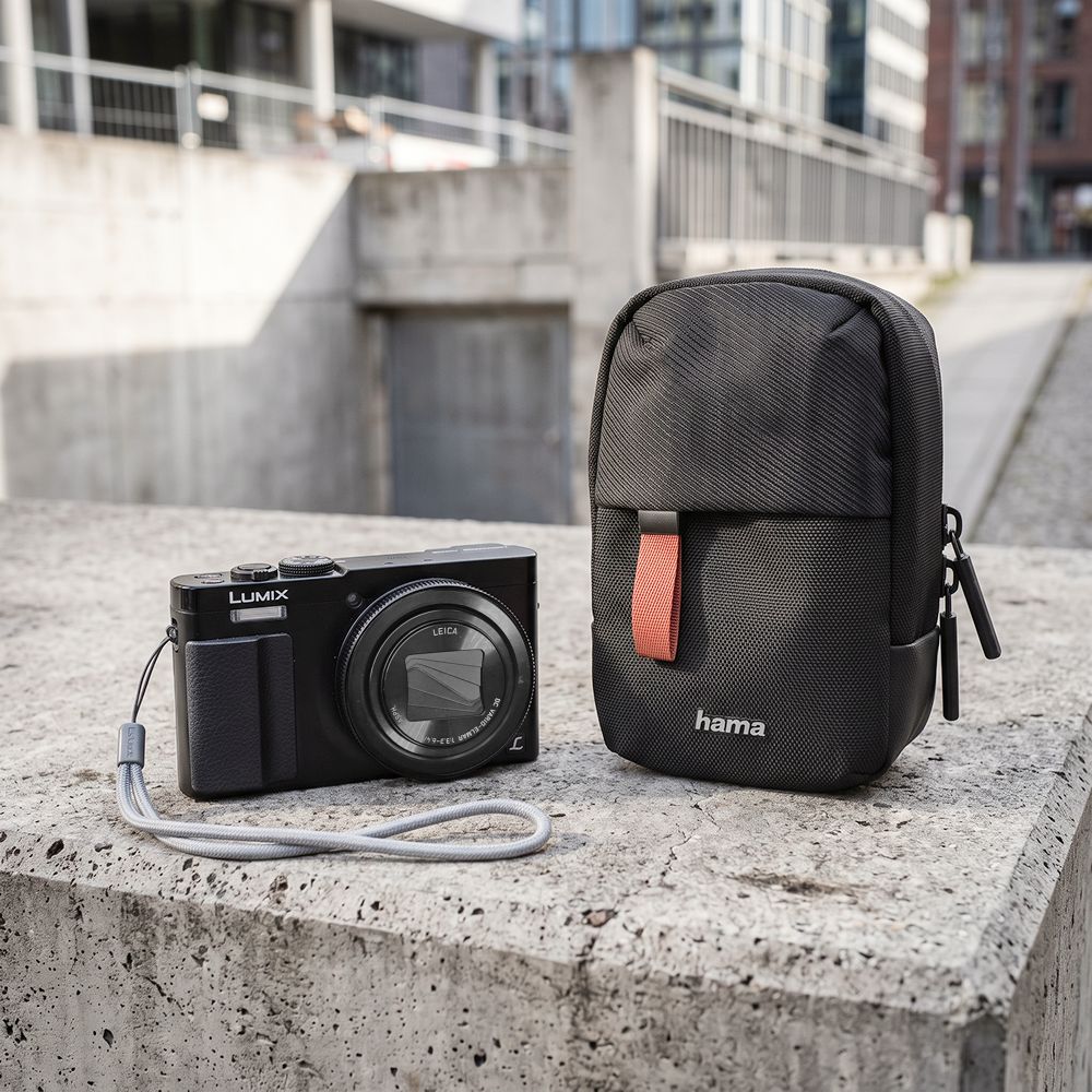 Schwarze kompakte Hama Fototasche mit Gürtelschlaufe auf einem Betonsockel neben einer Panasonic Lumix DMC-TZ71 Kamera, ideal auch für Panasonic Lumix DC-TZ202.