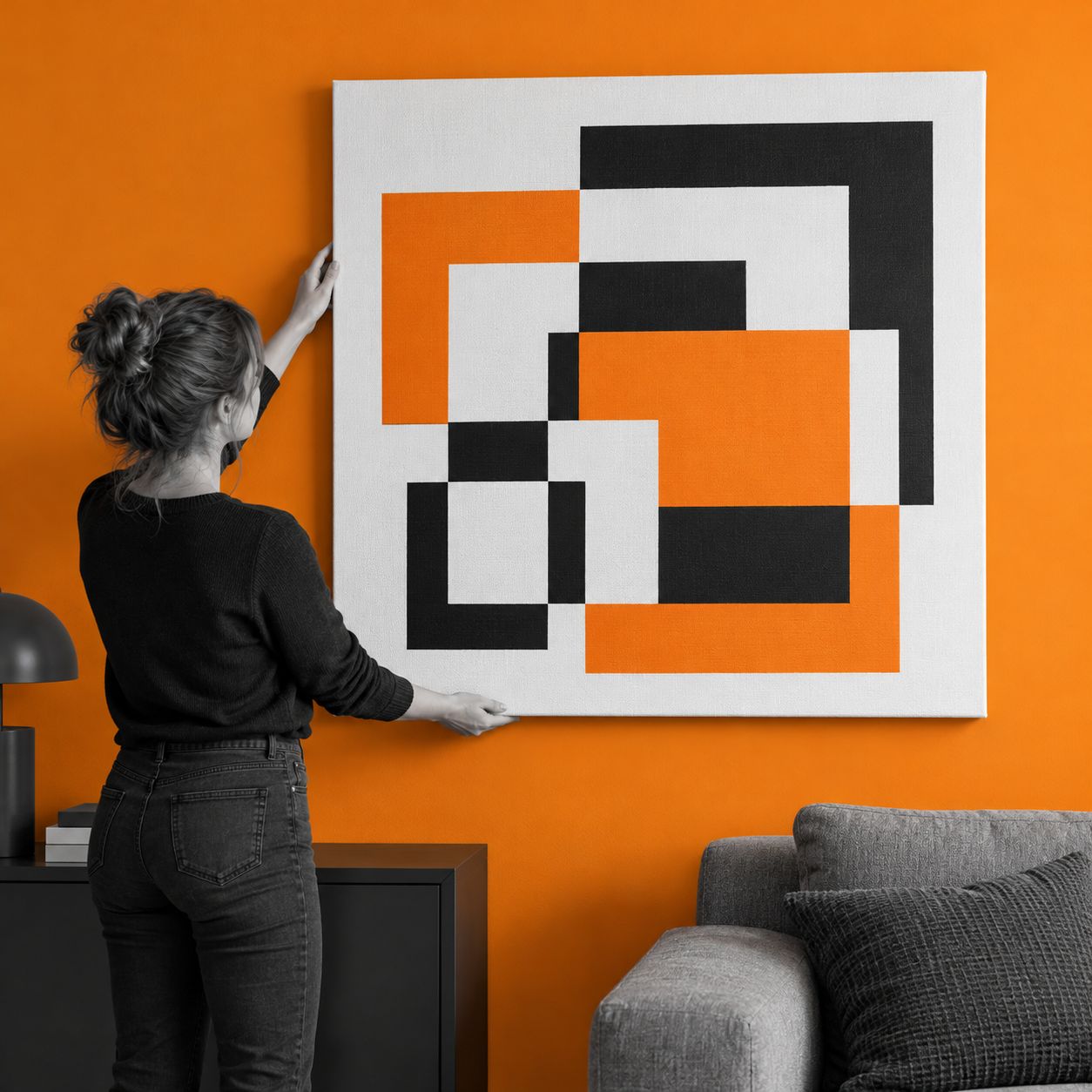 Ženska postavlja veliko kvadratno potiskano platno 100 × 100 cm ob oranžno steno nad sivo sedežno garnituro.