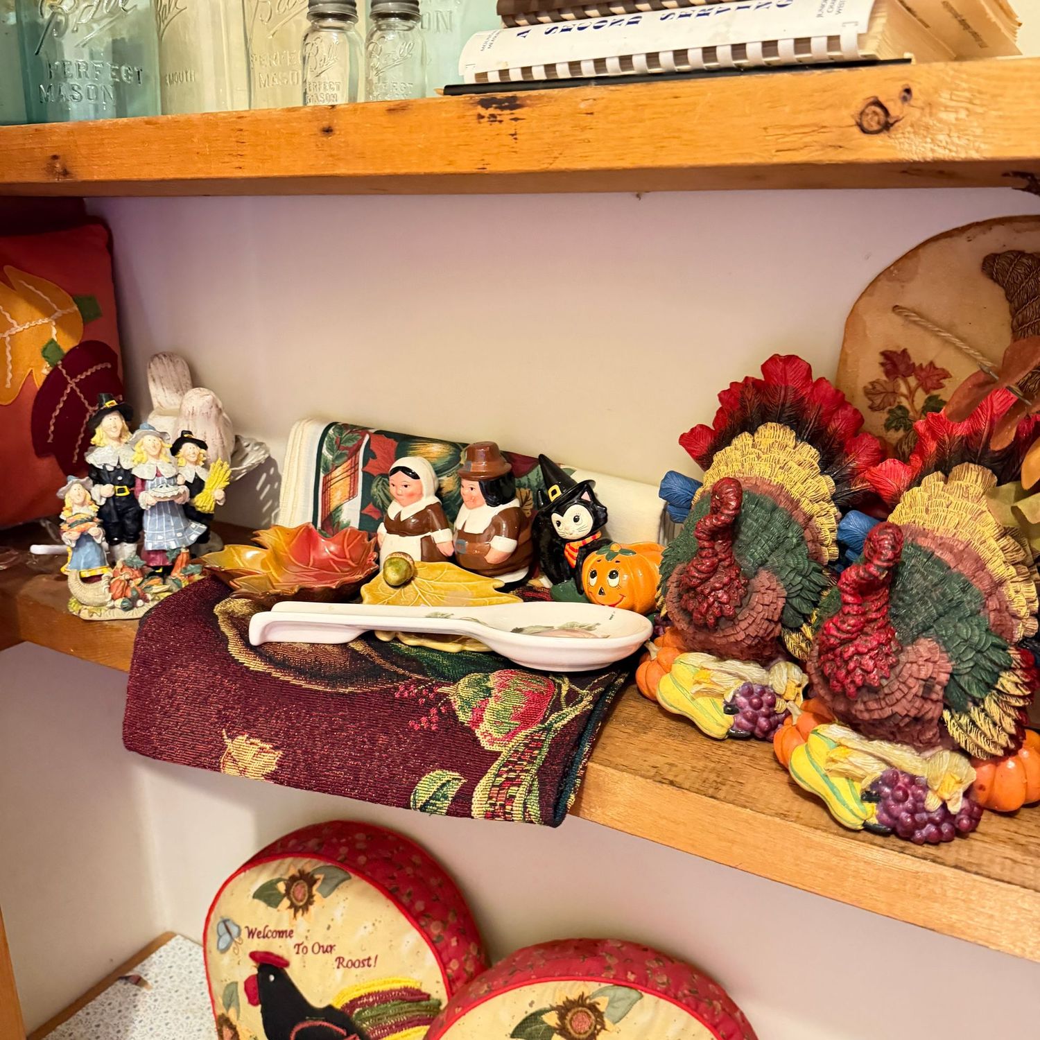 Shelf of fall decorations, including small pillow, figurines, spoon rest, table runners, resin 6"H turkeys, and cornucopia wall plaque, and fall flower arrangement 10"H