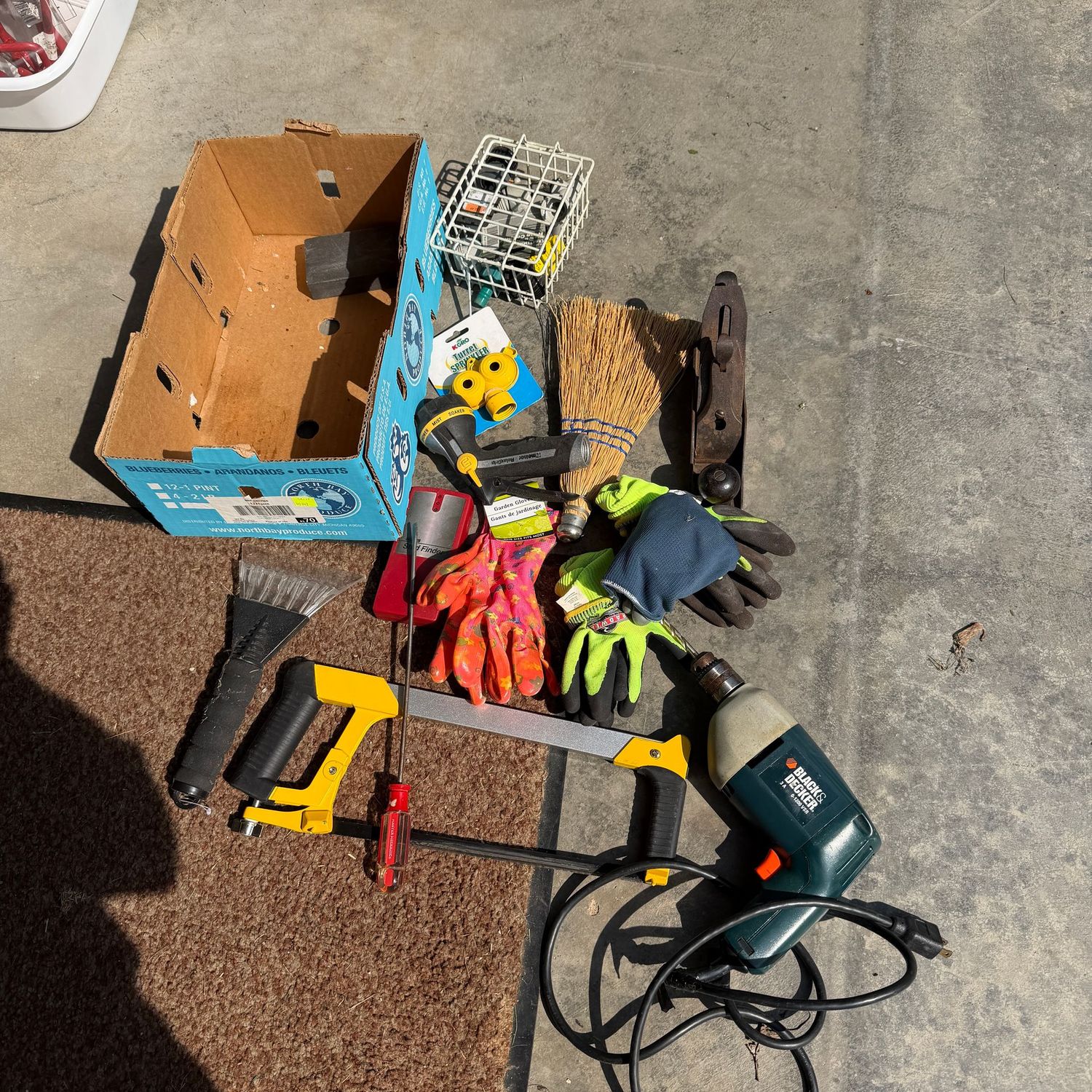 Small cardboard box with ice scraper, hand saw, corded drill, planer, work/garden gloves, wisk broom, sprinkler nozzles, sprayer