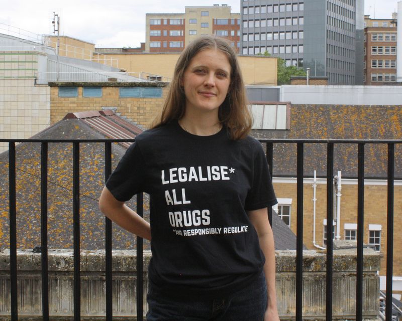 Ester is wearing a short-sleeved black t-shirt with ‘Legalise* all drugs *and responsibly regulate’ printed in white across the front.