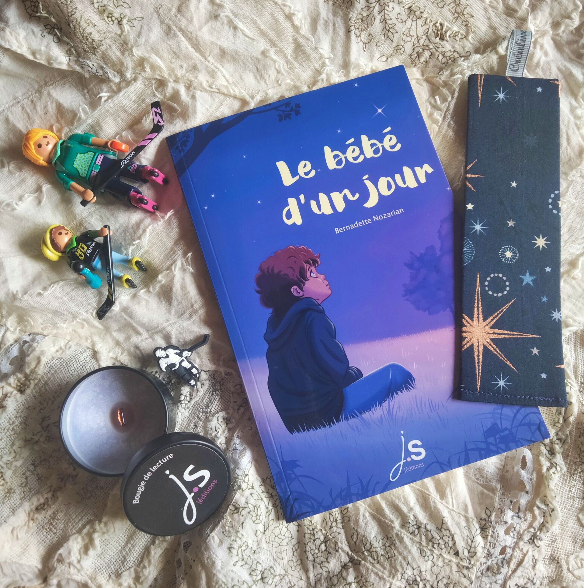 La couverture du bébé d’un jour, de Bernadette Nozarian est dans des tons bleu et violet. C’est une illustration de Layla Benabid qui présente une scène nocturne : un enfant blanc aux courts cheveux châtains est assis dans l’herbe à la campagne, dans un champ. On le voit de côté, il est tourné vers la droite. En haut à gauche apparaissent quelques branches d’un arbre. En arrière-plan devant l’enfant on voit deux silhouettes d’arbres. L’enfant a les yeux levés vers le ciel dans lequel brillent quelques étoiles dont une, particulièrement fort. Au premier-plan en bas de la couverture on voit quelques hautes herbes en plus gros plan. Au centre est placé le logo de la maison d’édition : JS Éditions en blanc et rose-violet pâle. Le titre est écrit en blanc dans une police manuscrite dans le ciel entre l’enfant et l’étoile la plus brillante sur 2 lignes. Le nom de l’autrice apparaît en plus petit sur une ligne dans une police sans serif en dessous.
