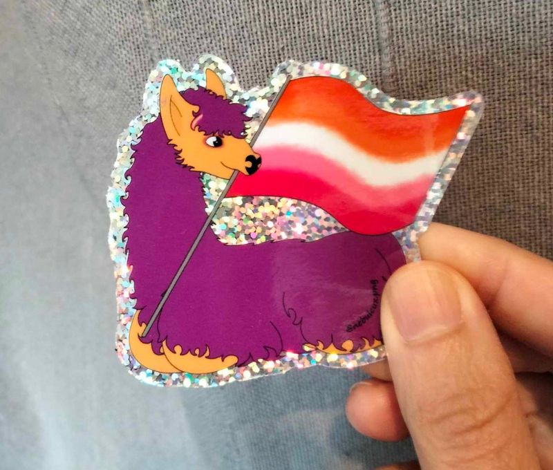 sticker découper à la forme de Barbouille le lama violet allongé et tenant dans sa bouche le drapeau lesbien. Il est pailleté. Il est signé Eli l'un des stagiaires de la maison d'édition