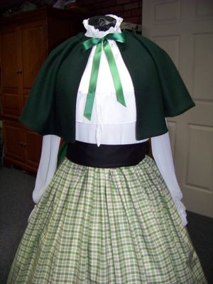 Christmas Caroler cape Hunter green solid wool blend cape reversible with black on inside one size fits most with black, green and beige plaid cotton homespun skirt and black sash. Handmade USA.