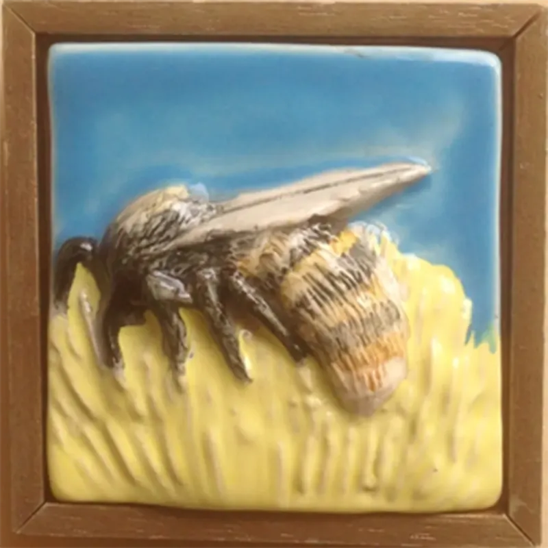 A framed cornflower blue 7cm x 7 cm tile with an under-glazed orange flower and yellow bee 8.5cm x 8.5cm with frame.