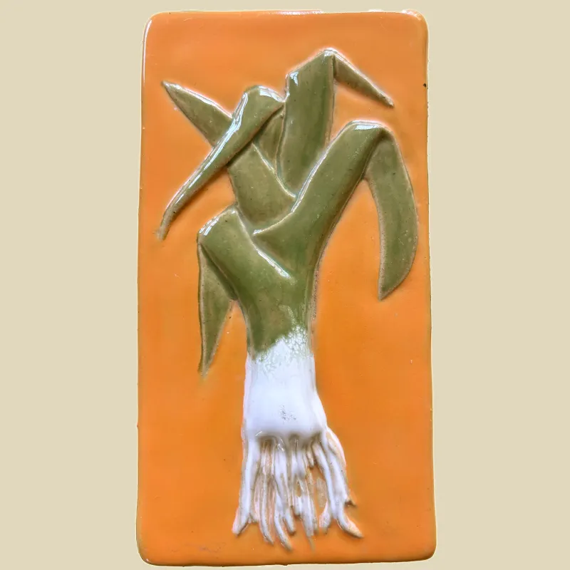 This is an unframed ceramic feature tile titled "Leek" It is hand painted in orange and green glazes