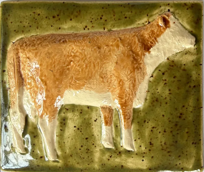 A feature tile depicting a hereford cow on a mottled green background 14cm x 11.75cm
