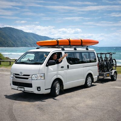 White Toyota HiAce with kayak and bikes at New Zealand beach carpark, outdoor adventure scene
