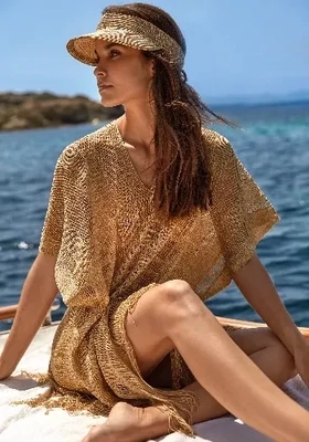 Tessy Indira Jany Golden Mesh Dress. The fabric is a gold mesh design. The sleeves are half length and there is fringing at the hem and V-neck collar. Lifestyle photo.