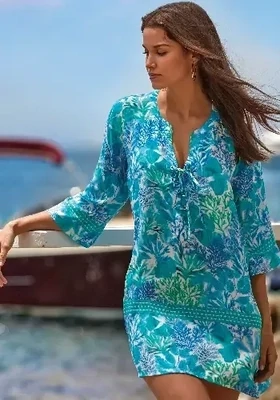 Roidal Tessy Coral Suen Tunic Dress. The fabric is a turquoise blue and sea green design. The dress has a a deep notch v-neckline and ¾ length sleeves, the hemline falls above the knee. Lifestyle photo.