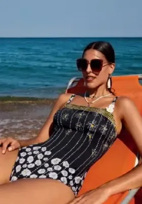 Magda Shaper Swimsuit by Nuria Ferrer Magda Shaper Swimsuit by Nuria Ferrer. The black suit features stylised drawings daisies joined by chains of dots, and some of the pretty golden yellow daisy centres are softly heart shaped. Lifestyle photo.