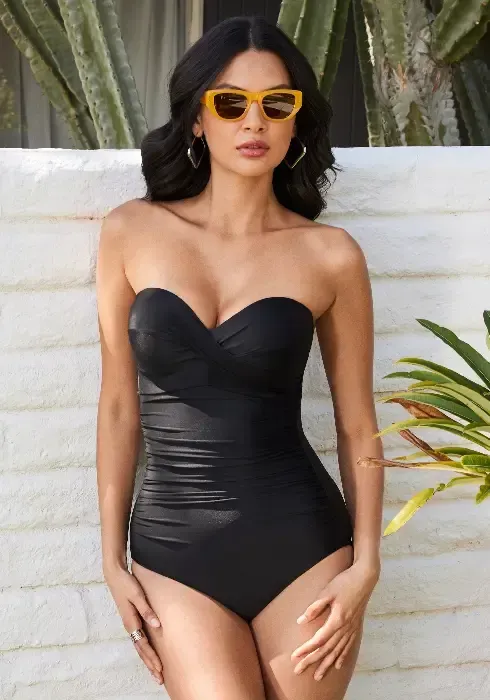 Madrid Black Bandeau Swimsuit by Miraclesuit. A classic bandeau style swimsuit with the addition of completely removable shoulder straps. Lifestyle photo with model wearing sunglasses.