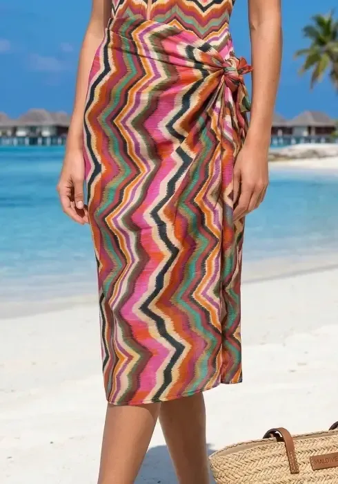 A model wears a colourful chevron-print sarong tied at the waist on a sunny tropical beach with matching swimsuit.