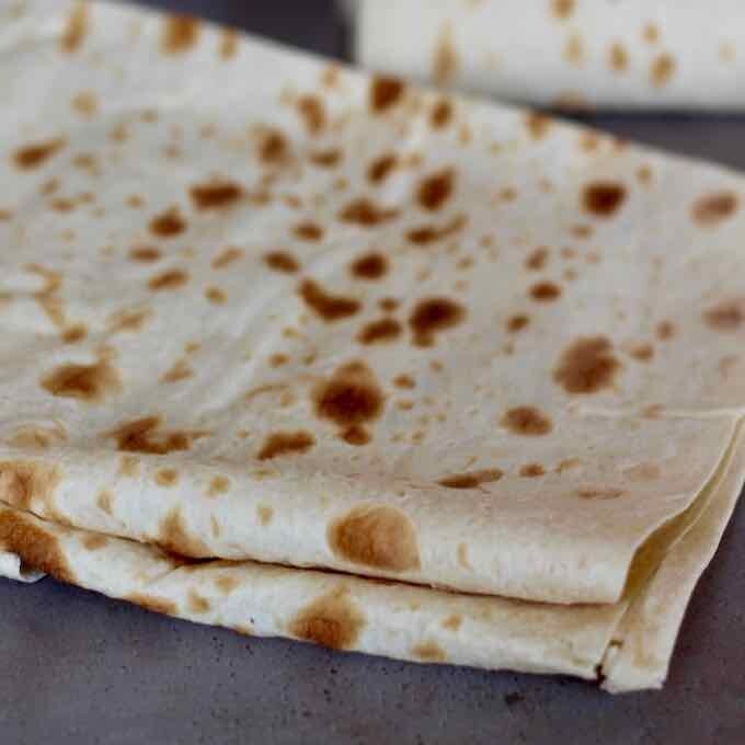 "Lavash bread, a thin and soft flatbread, is laid out on a wooden board. Its surface is smooth with occasional golden-brown spots, highlighting its light and airy texture.