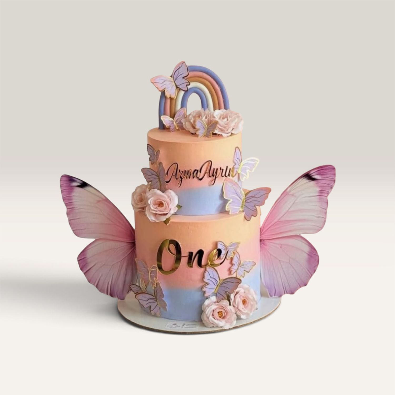 Torta de mariposas de dos pisos con arcoíris, flores rosadas, alas grandes y tonos pastel.