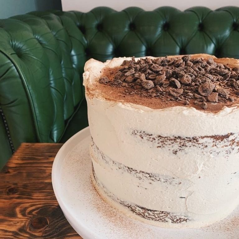 Tiramisu Cake - A cake version of the beloved Italian dessert — light, creamy, and full of flavour. Three layers of espresso-soaked sponge, iced in a semi-naked finish with whipped vanilla mascarpone cream, and topped with a generous dusting of dark chocolate shavings. Simple, elegant, and irresistibly good.