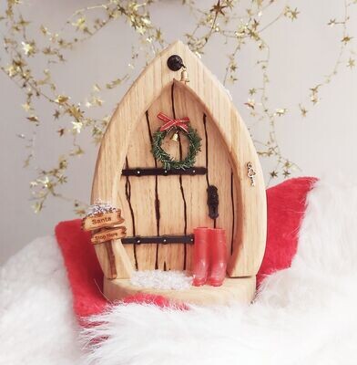 An oak fairy door sat on a Santa hat featuring  a Christmas wreath, red wellies, snow and a santa stop here sign.  
