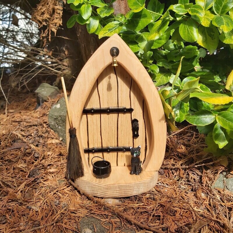 A fairy door sat on the forest floor.  The door has a broom, cauldron and a black cat on the doorstep plus a small gold bell.