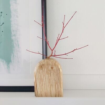 A small wooden vase on a white shelf with a black framed blue picture in the background.  The vase has red twigs in it.
