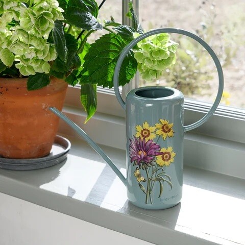Asteraceae Design.  Light Duck Egg Blue with three yellow flowers and one purple flower design.
Image of indoor watering can with long cylindrical water holder long thing pouring spout and large tin circular handle.
Lifestyle image of watering can placed on a windowsill next to a pot plant.