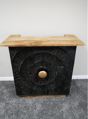 A very useful addition to your home. Its a bar counter that doesn't take up too much space. At just 114 cm wide, you will wonder how you did without it. This image shows the counter and the attractive front.
It is made from mango wood, and has a interesting circular design on the black front of the counter. It's just the right height too, when you are pouring the drinks, and it sports very useful shelves on the reverse side for storing glasses or booze!  