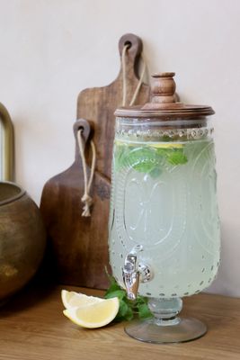 Clear glass drink dispenser with decorative embossed detailing, a wooden lid with turned knob, metal spigot, & pedestal base