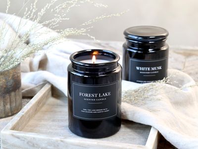 Lucon Scented Candle - White Musk - White Candle in Dark Glass Container.  Black Label with White Writing.  Shown Lit