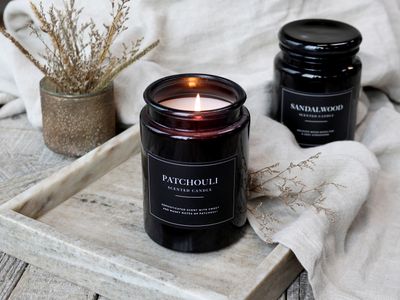 Lucon Scented Candle - Patchouli - White Candle in Dark Glass Container.  Black Label with White Writing.  Shown Lit