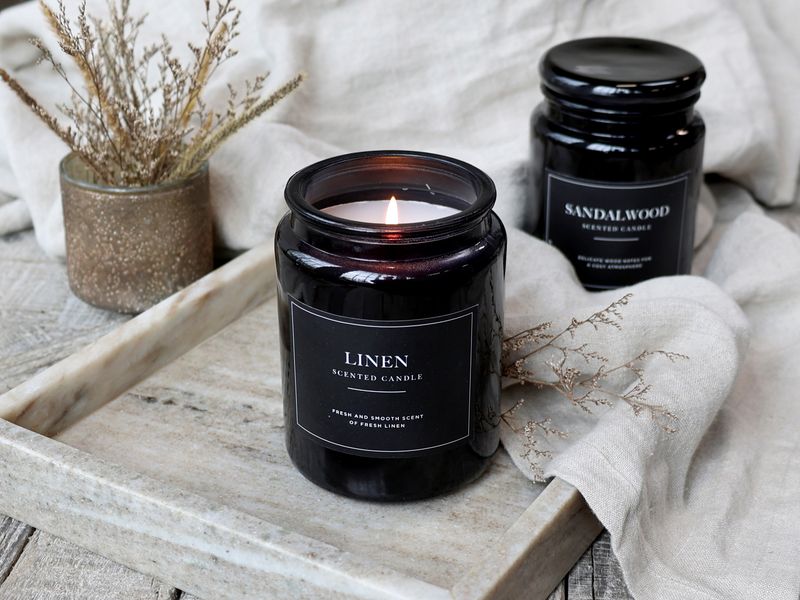 Lucon Scented Candle - Linen - White Candle in Dark Glass Container. Black Label with White Writing. Shown Lit