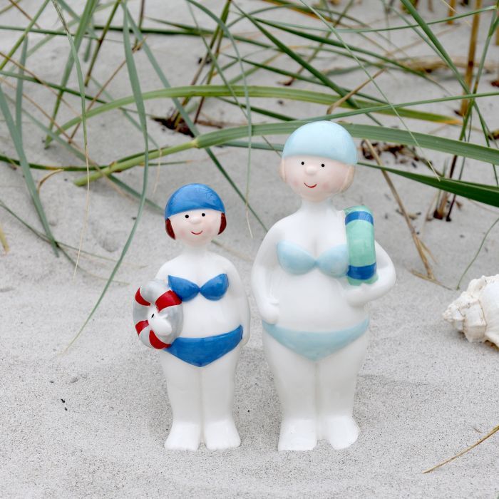 Two cheerful beach-themed figurines in swimsuits standing on sand, holding a lifebuoy and beach towel by dune grass.