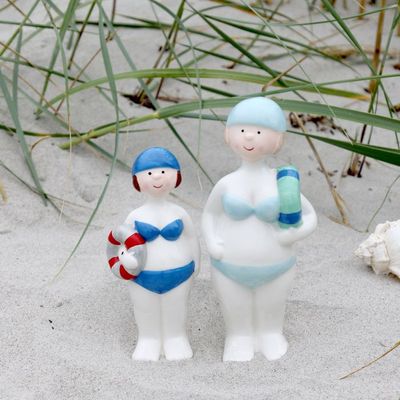 Two cheerful beach-themed figurines in swimsuits standing on sand, holding a lifebuoy and beach towel by dune grass.