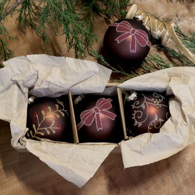 Three mocha Christmas baubles decorated with glittering bows, botanical sprigs and scrolls, arranged among evergreen foliage.