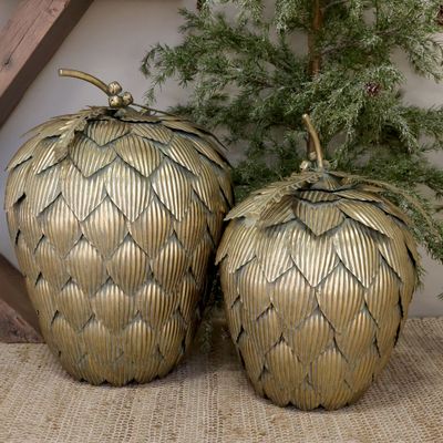 Two large gilded artichoke Christmas decorations with layered ribbed leaves, displayed beside a frosted evergreen tree.