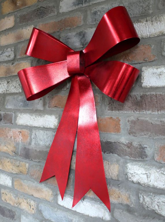 Large sculptural scarlet Christmas bow with a metallic sheen and long ribbon tails, displayed on a brick wall.