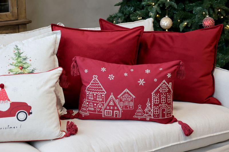 Scarlet rectangular cushion with a cream gingerbread village, Christmas trees, snowflakes and tassels at each corner.
