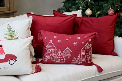Scarlet rectangular cushion with a cream gingerbread village, Christmas trees, snowflakes and tassels at each corner.
