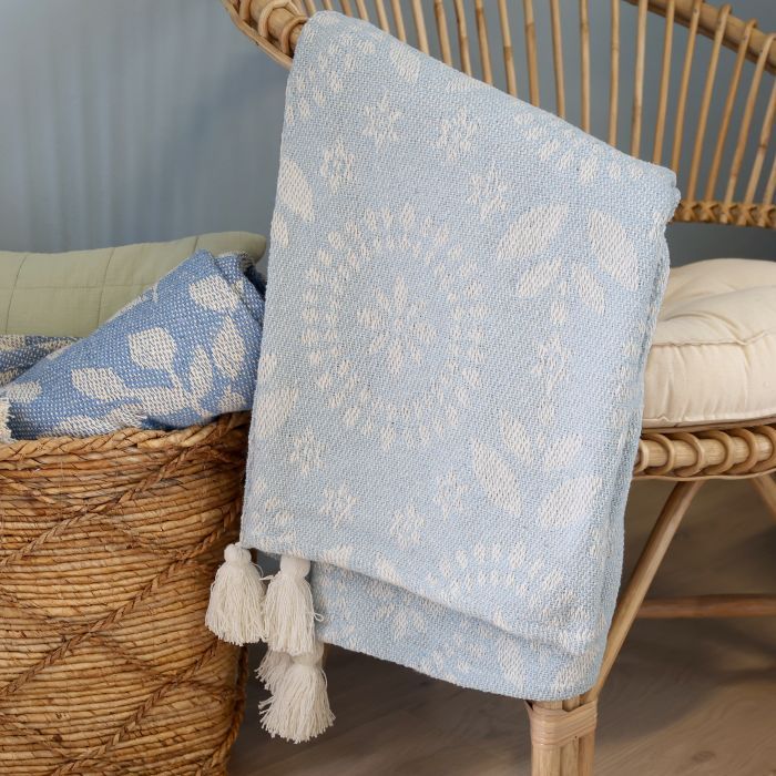 Soft blue woven throw with delicate ivory floral detailing, textured finish and decorative cream tassels at the corners.